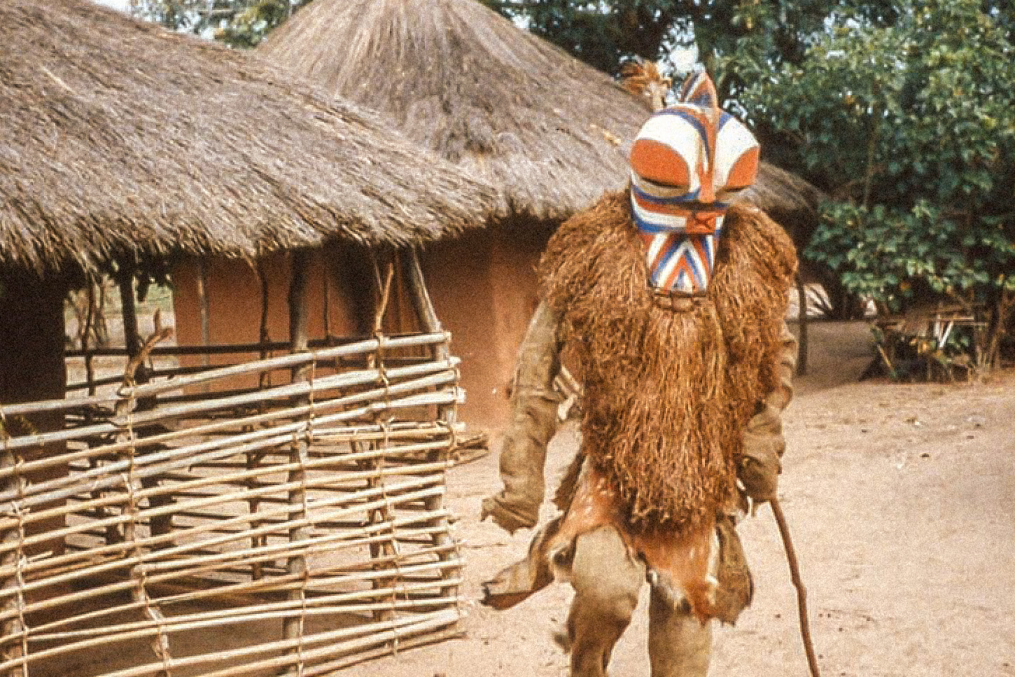 Masker with blue, orange, and white facial striations and a costume made of burlap. kalebwe, Ebombo chiefdom, kalongo. Photo: karEL PLaSMaNS, 1960S; © DuNja hErSak